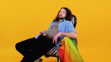 Gökkuşağı çizgili bayraklı 20 'li yaşlarda genç beyaz bir adam sarı arka planda izole bir sandalyede oturuyor. Insanlar yaşam tarzı lgbtq konsepti.
