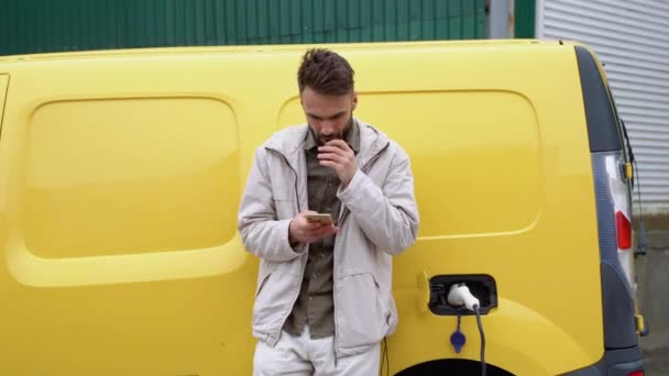 Junger Mann Raucht Eine Elektronische Zigarette Während Elektroauto Außenstation Auflädt — Stockvideo