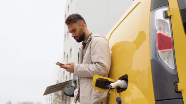 電気自動車充電ステーションで車を充電しながらスマートフォンを持っている男 クローズアップ — ストック動画