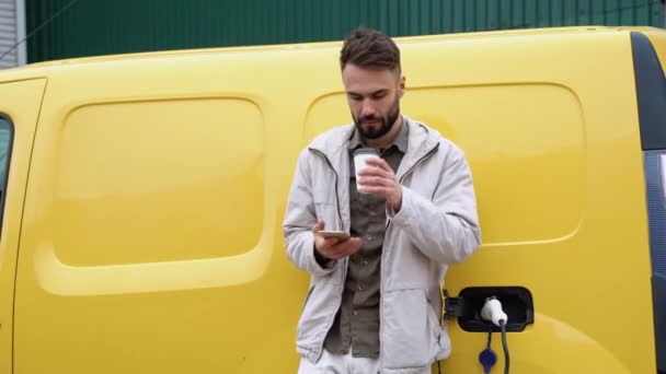 現代の電気自動車の近くに立つスタイリッシュなハンサムな男性とスマートフォンの屋外でのテキストメッセージ — ストック動画