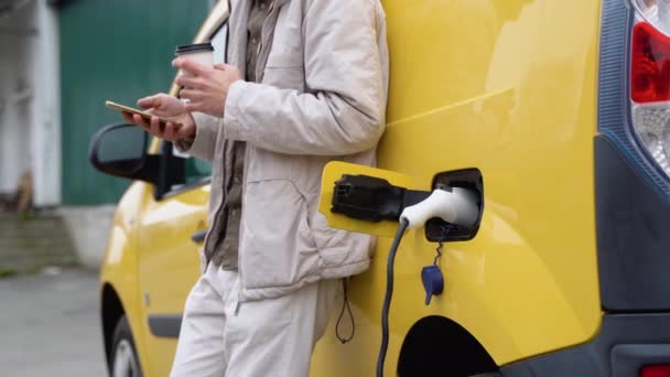 Junger Mann Chattet Mit Handy Während Elektroauto Außenstation Auflädt — Stockvideo