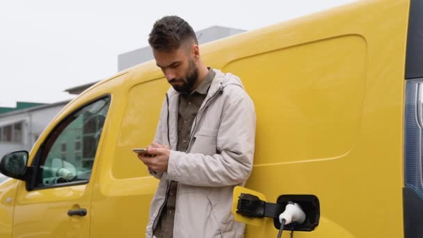 現代の電気自動車の近くに立って スマートフォンの屋外でテキストメッセージ幸せなハンサムな男性 携帯電話で良い気分でブラウジングの若い男 都市概念 — ストック動画