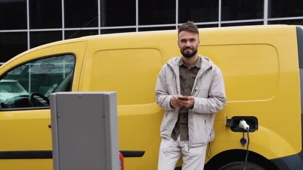 Uśmiechnięty Mężczyzna Swobodnym Ubraniu Telefonem Opierający Się Samochód Elektryczny Stojący — Wideo stockowe