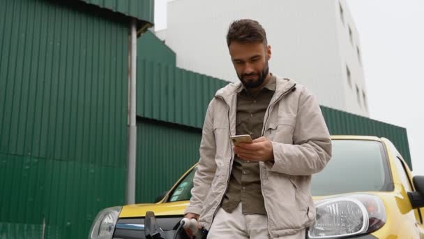 現代の電気自動車の近くに立つスタイリッシュなハンサムな男性とスマートフォンの屋外でのテキストメッセージ 携帯電話で良い気分のブラウジングの若い男 — ストック動画