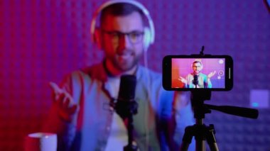 Online podcast sırasında kameraya bakarak konuşan beyaz blogcu akıllı telefon kaydına odaklanmış bir çekim. İçerik yaratıcı çevrimiçi yayın yapıyor, blog yazarı kulaklık takmayı tartışıyor.