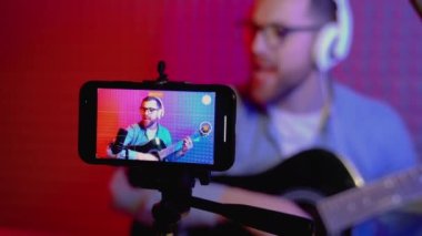 Podcast sırasında gitar çalan genç bir etken veya evdeki cep telefonundan izleyiciler için canlı video yayını - sanat, hobi ve video blogu kavramı.
