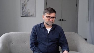 Battaniyeye sarılı yaşlı bir adam kanepede otururken burun spreyi sıkar..