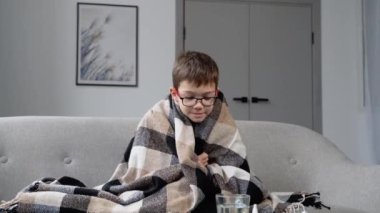 Grip, ateş ve baş ağrısı olan küçük bir çocuk kanepede ekoseye sarılı oturuyor. Mevsimsel hastalıklar dönemi. Viral hastalıklar kavramı.