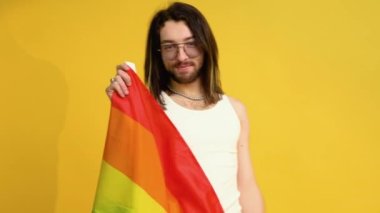 Genç aktivist gülümseyen neşeli gay adam sarı arka plan stüdyosunda izole edilmiş gökkuşağı bayrağı sallıyor. İnsanlar yaşam tarzı kavramını seviyorlar.