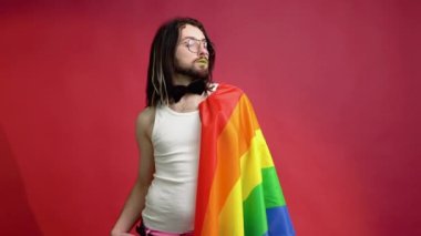 Genç aktivist gülümseyen mutlu gay adam kırmızı arka plan stüdyosunda izole edilmiş gökkuşağı bayrağı tutuyor.