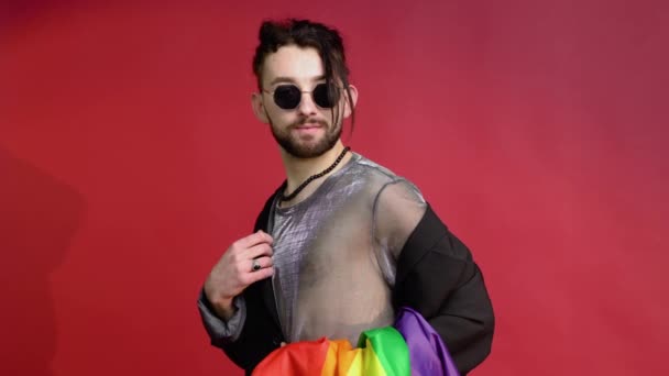 メッシュのTシャツとサングラスを身に着けているゲイの男性は 多色の虹の旗で赤い背景に座っています 概念の多様性 — ストック動画