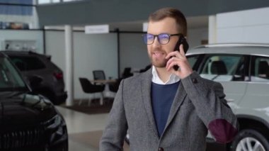 Gözlüklü ve takım elbiseli beyaz adam araba galerisinde telefonla konuşuyor..