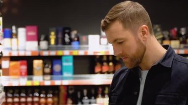 Bir adam süpermarketin rafından alkollü içecekler alır. Alkol almak için markete gidiyorum..