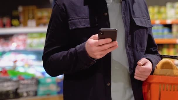 携帯電話で購入のリストを見てスーパーマーケットに立っている若い男の作物のイメージ — ストック動画