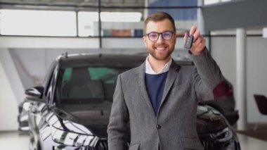 Portrait of happy adult successful man posing in auto showroom buying new automobile. Positive male smiling for camera and demonstrating keys while standing near new vehicle in showroom