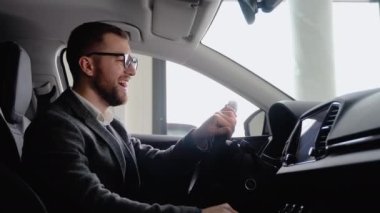 Happy man sits in new electric car in shop dealership and celebrate purchase of new vehicle. Buy car.