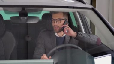 Gözlüklü ve takım elbiseli beyaz bir adam yeni bir arabada oturuyor ve bir araba fuarında telefonla konuşuyor.