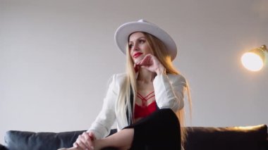 Boyalı saçlı, şapkalı, makyajlı beyaz ceketli ve kırmızı sutyenli bir kadın stüdyoda poz veriyor. Moda çekim konsepti.