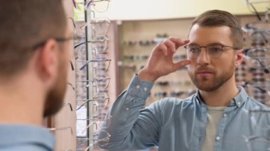 Portrait of handsome bearded guy picking new specs at optical shop, looking at mirror. Choosing glasses concept.