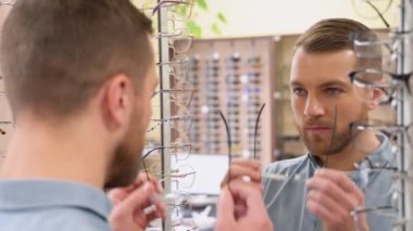 Portrait of handsome bearded guy picking new specs at optical shop, looking at mirror. Choosing glasses concept