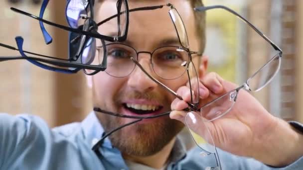 興奮した男は光学店で彼の手の中に多くの新しいメガネを保持しています メガネのコンセプトを選ぶ — ストック動画