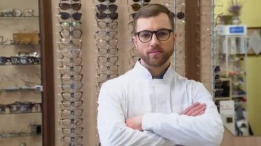 Attractive young doctor in ophthalmology clinic. Doctor ophthalmologist is standing near shelves with different eyeglasses.