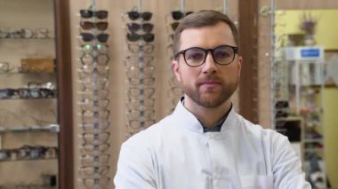 Attractive young doctor in ophthalmology clinic. Doctor ophthalmologist is standing near shelves with different eyeglasses.