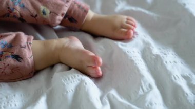 Sevimli masum bebek ilk bebek emeklediğinde yatakta emeklemeye çalışıyor. Güzel bebek kız pembe bebek elbisesi giyiyor. Küçük çocuk bacağını ve vücudunu kaldırmak için kullanmaya çalışıyor. Bacaklarını kapat..