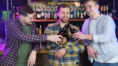 Handsome friends clinking bottles of beer and smiling while resting at the pub.