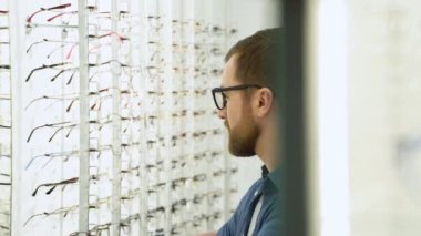 Portrait of handsome bearded guy picking new glasses at optical shop, looking at mirror