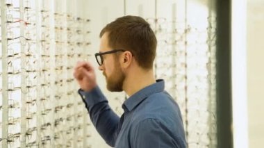 Portrait of handsome bearded guy picking new glasses at optical shop, looking at mirror