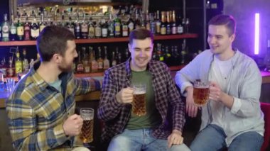 Group of happy young men drinking cold draft beer, chatting and having good time at pub. Smiling carefree friends enjoying drinking together in bar.