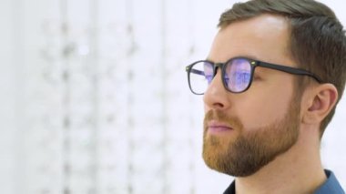 Portrait of handsome bearded guy picking new specs at optical shop, looking at mirror