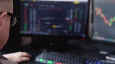 Trader wearing eyeglasses sitting at desk at office monitoring stock market looking at monitors analyzing candle bar price flow touching chin concerned trading concept