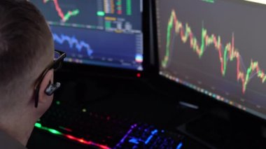 Borsa borsası. Modern ofisinde çalışırken bilgisayar ekranındaki verileri kalemle işaret eden ve bir elinde Bitcoin tutan gözlüklü genç profesyonel tüccarın geri görünümü.