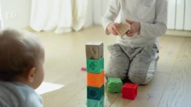 Two childs building tower of block toys sitting on floor in sunny bedroom.