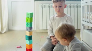 Two childs building tower of block toys sitting on floor in sunny bedroom.