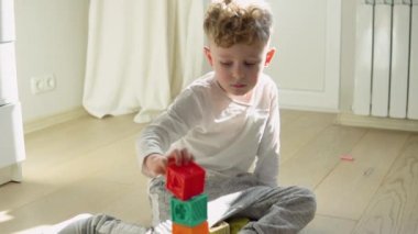 Little boy building tower of block toys sitting on floor in sunny bedroom.