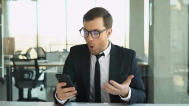 Angry boss shouts to the employee while looking at phone, dismisses the subordinate, a businessman in a business suit sits at a table in a modern office.