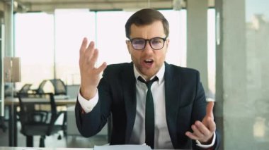 Angry boss shouts at the camera to the employee, dismisses the subordinate, a businessman in a business suit sits at a table in a modern office.