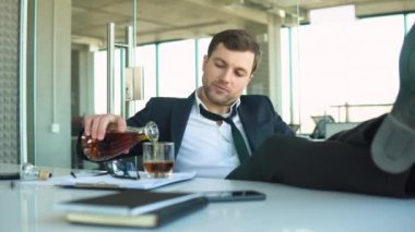 Things getting worse, difficulties at job. Frustrated young businessman drinking cause of having stress, holding glass of whiskey, holding his head, office interior