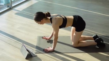 Beautiful sporty woman in sportswear doing plank with leg and hand raise while watching training video on tablet. Indoor studio shot illuminated by sunlight from window