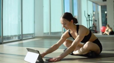 Bun saç stili ve dar spor giyimli mutlu güzel bir kadın tabletten eğitim videosu izlerken bacak yükseltme tahtası yapıyor. Pencereden gelen güneş ışığıyla aydınlatılan kapalı bir stüdyo..