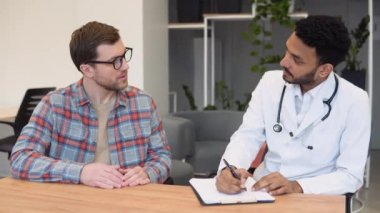 Hintli erkek doktor konsültasyonda hastaya danışmanlık yapıyor. Profesyonel doktor beyaz önlüklü yetişkin bir adamla konuşuyor. Klinikteki randevuda tıbbi evrakları imzalıyor..