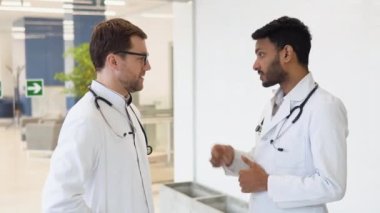 Medical multiethnic staff having discussion in a hospital hallway. Indian and caucasian smiling doctors in white coat working in a medical clinic. Two hospital workers discussing on laboratory test