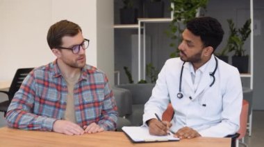 Hintli erkek doktor konsültasyonda hastaya danışmanlık yapıyor. Profesyonel doktor beyaz önlüklü yetişkin bir adamla konuşuyor. Klinikteki randevuda tıbbi evrakları imzalıyor..