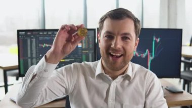 Satisfied man holding a golden bitcoin in his hand against the background of financial charts