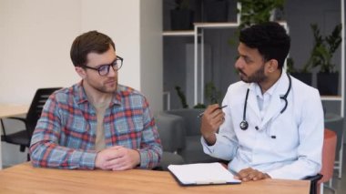 Indian male doctor consulting patient filling form at consultation. Professional physician wearing white coat talking to adult man signing medical paper at appointment visit in clinic.