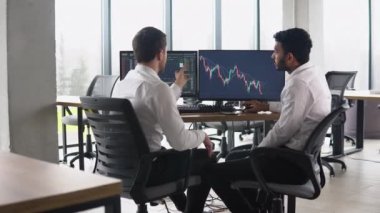 Two multiracial investors brokers analysing indexes online cryptocurrency stock. Diverse managers traders analysts discussing financial growth market at desk with pc graphs on screen.