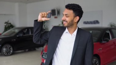 Indian salesman standing at dealership near new luxury car and demonstrating keys